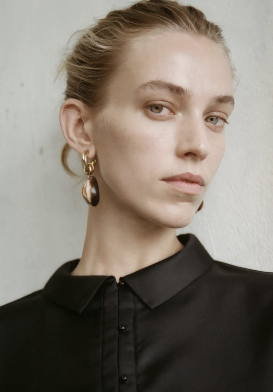Close-up of a fair-skinned woman with blonde hair, wearing a black collared shirt and stylish earrings against a neutral background.