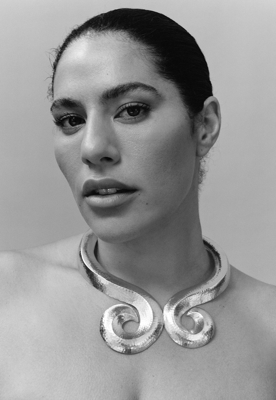 Black and white portrait of a woman with dark hair and a statement silver necklace, facing forward in a close-up studio shot.