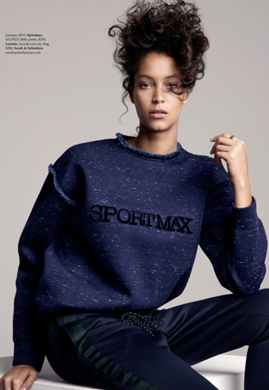 A woman with curly hair wears a Sportmax sweatshirt and pants while posing indoors with a neutral background in a medium shot.