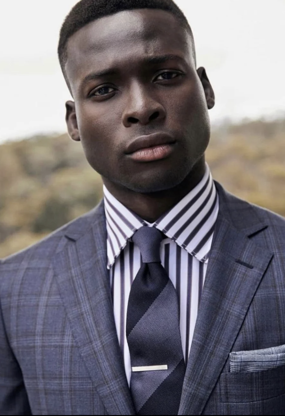 Headshot of a dark-skinned adult male in a tailored grey suit with a striped shirt and diagonally striped tie outdoors in soft lighting.