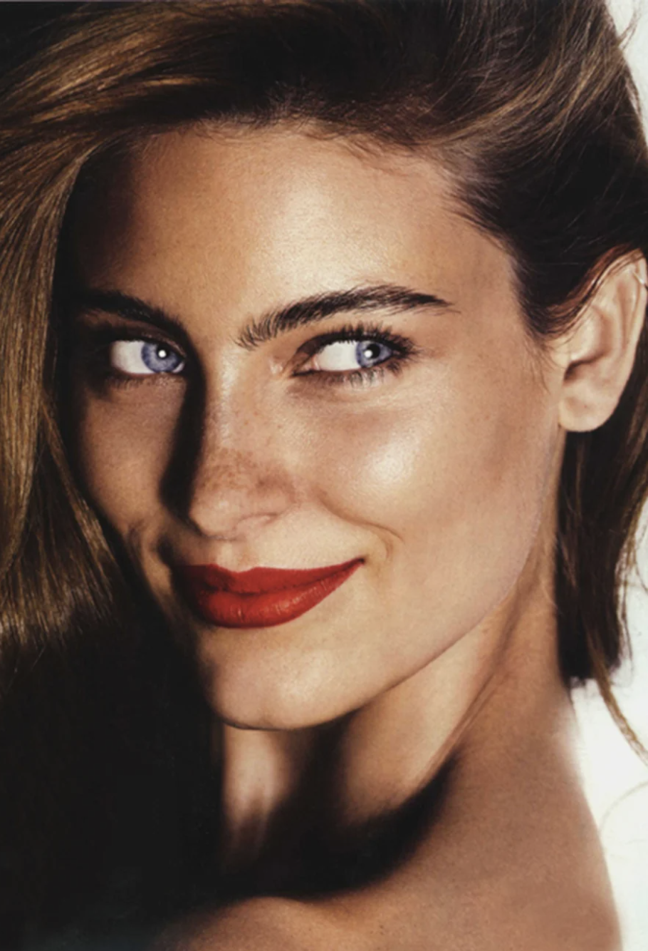 Close-up studio portrait of a smiling adult female model with blue eyes, light brown hair, red lipstick, fair skin, and freckles.