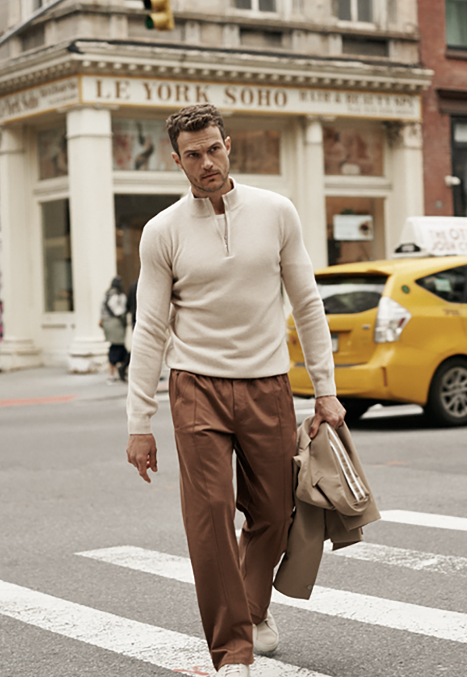 A stylish man walks across a crosswalk in an urban setting. He wears a cream-colored sweater and brown trousers while holding a jacket.