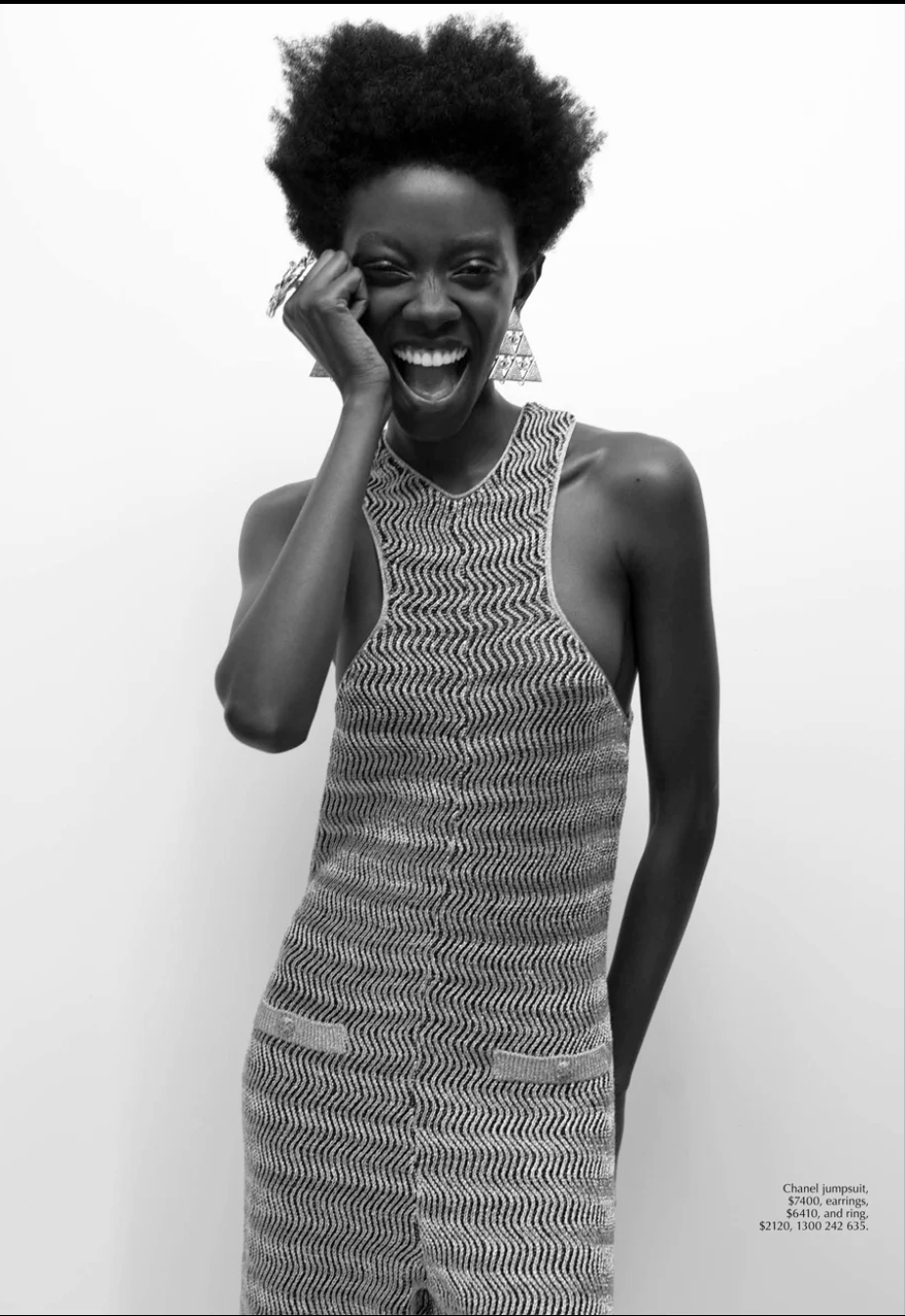 Black woman in stylish jumpsuit laughs, accessorized with large earrings and ring, set against a plain white background, black and white