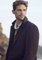 Close-up portrait of a male model in a dark blazer, striped polo shirt, and knitted scarf outdoors near water.