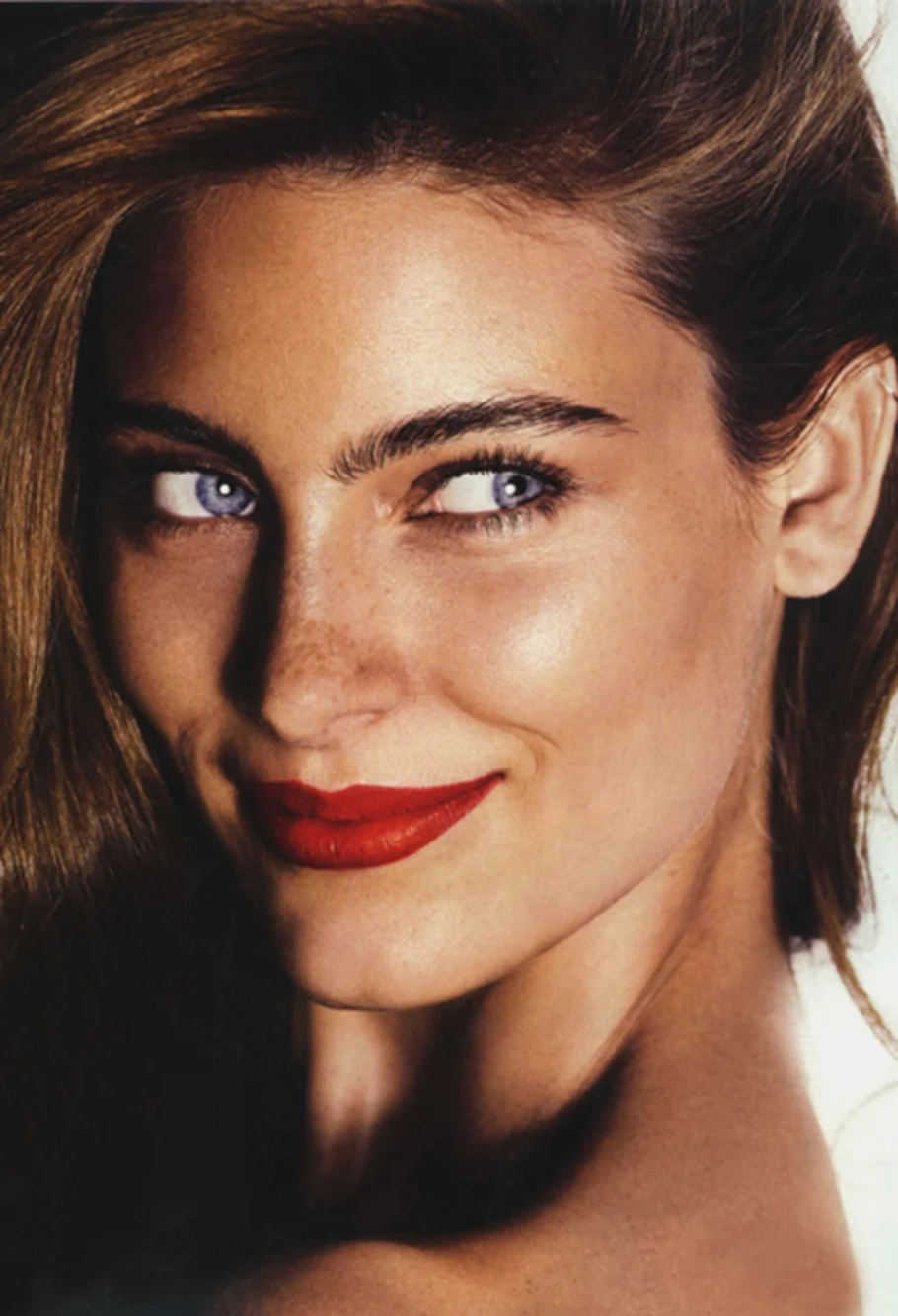 Close-up portrait of a woman with vibrant red lipstick, striking blue eyes, and light brown hair against a plain white backdrop.