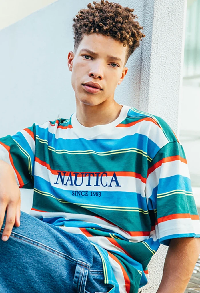 Young man with curly hair wearing a striped t-shirt leaning against a white textured wall