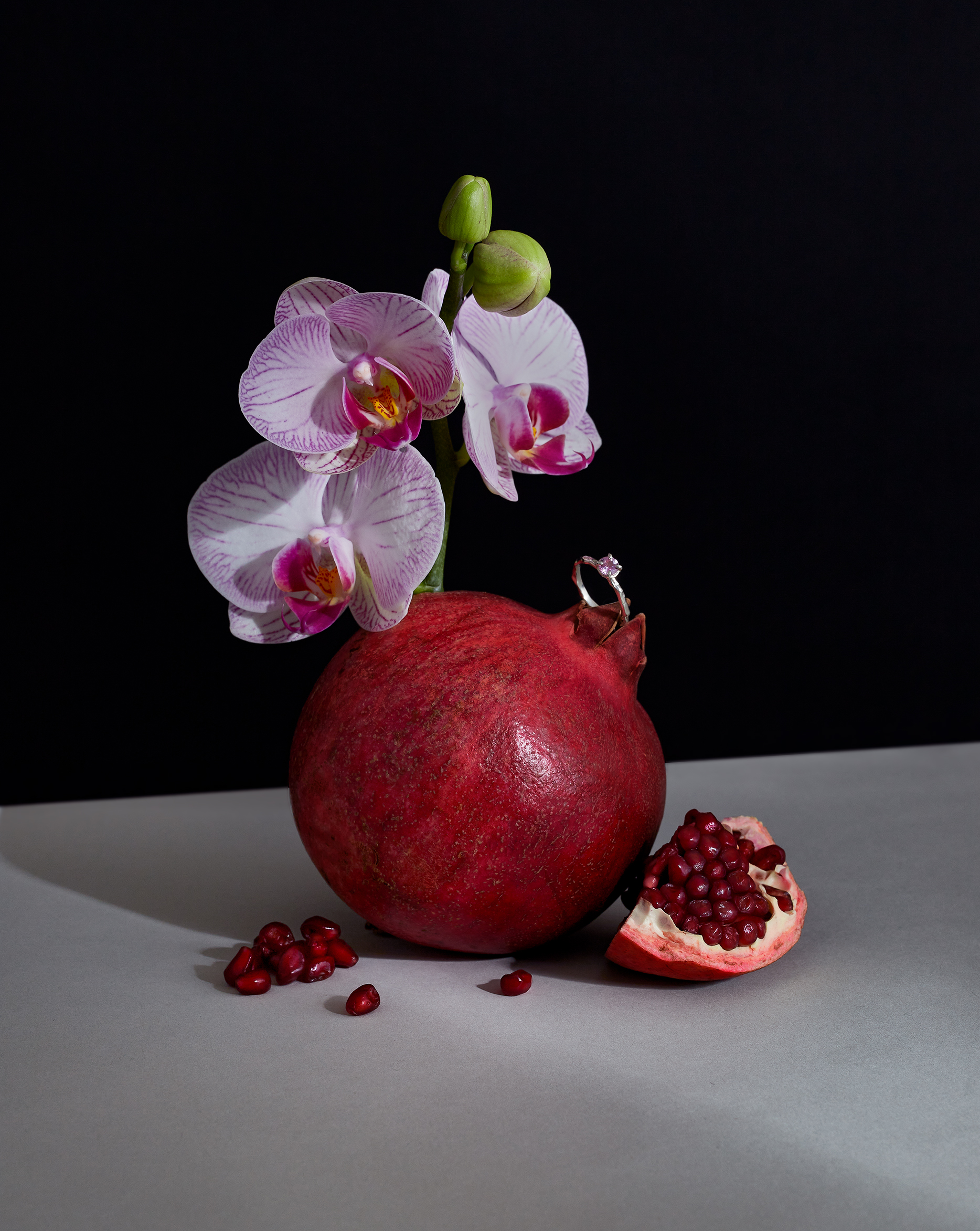 Still life featuring a vibrant red pomegranate topped with a delicate white orchid and a diamond ring, set against a dark backdrop.