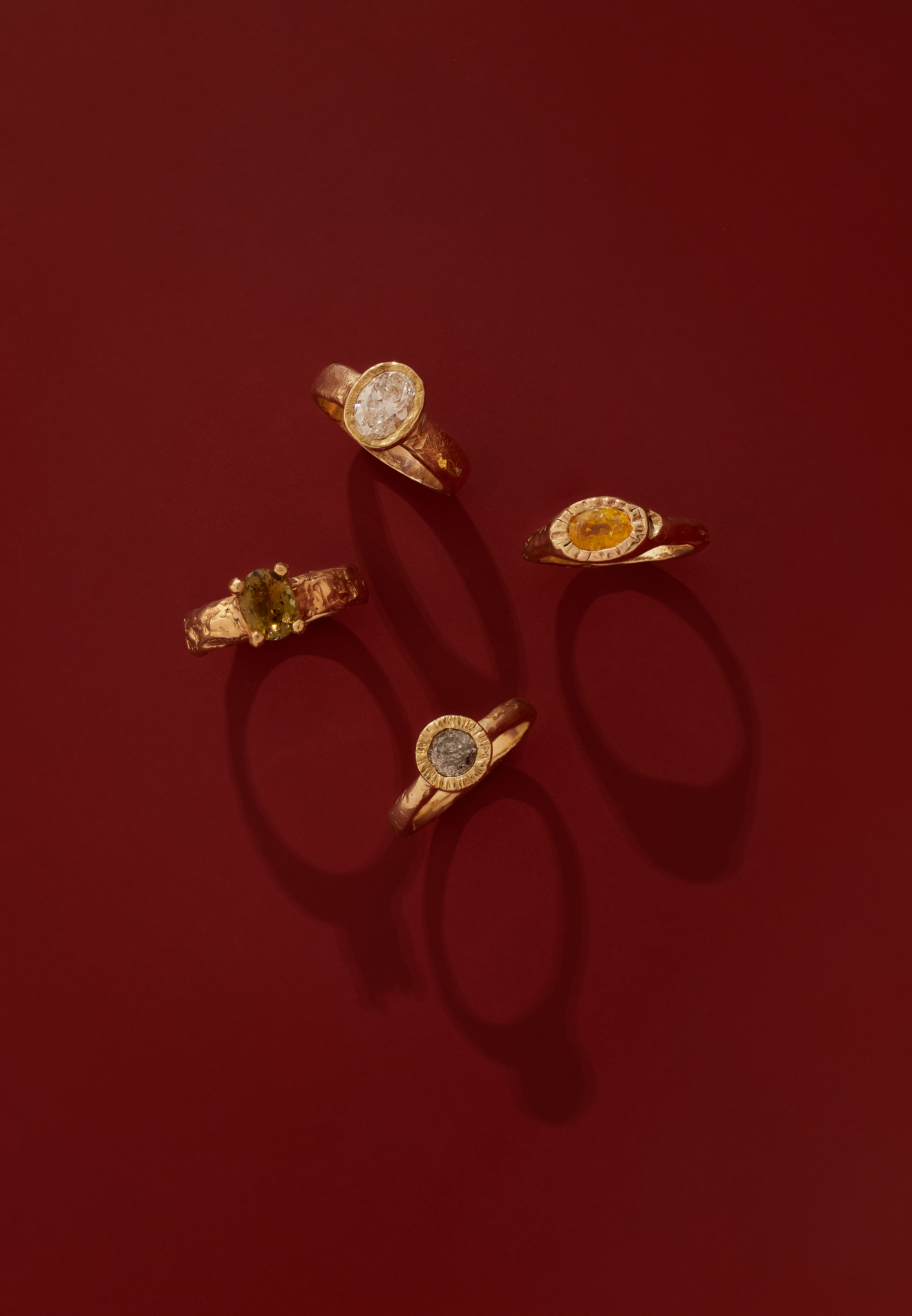 Four gold rings set with various gemstones are arranged on a red surface with soft shadows, in a high-angle product shot.