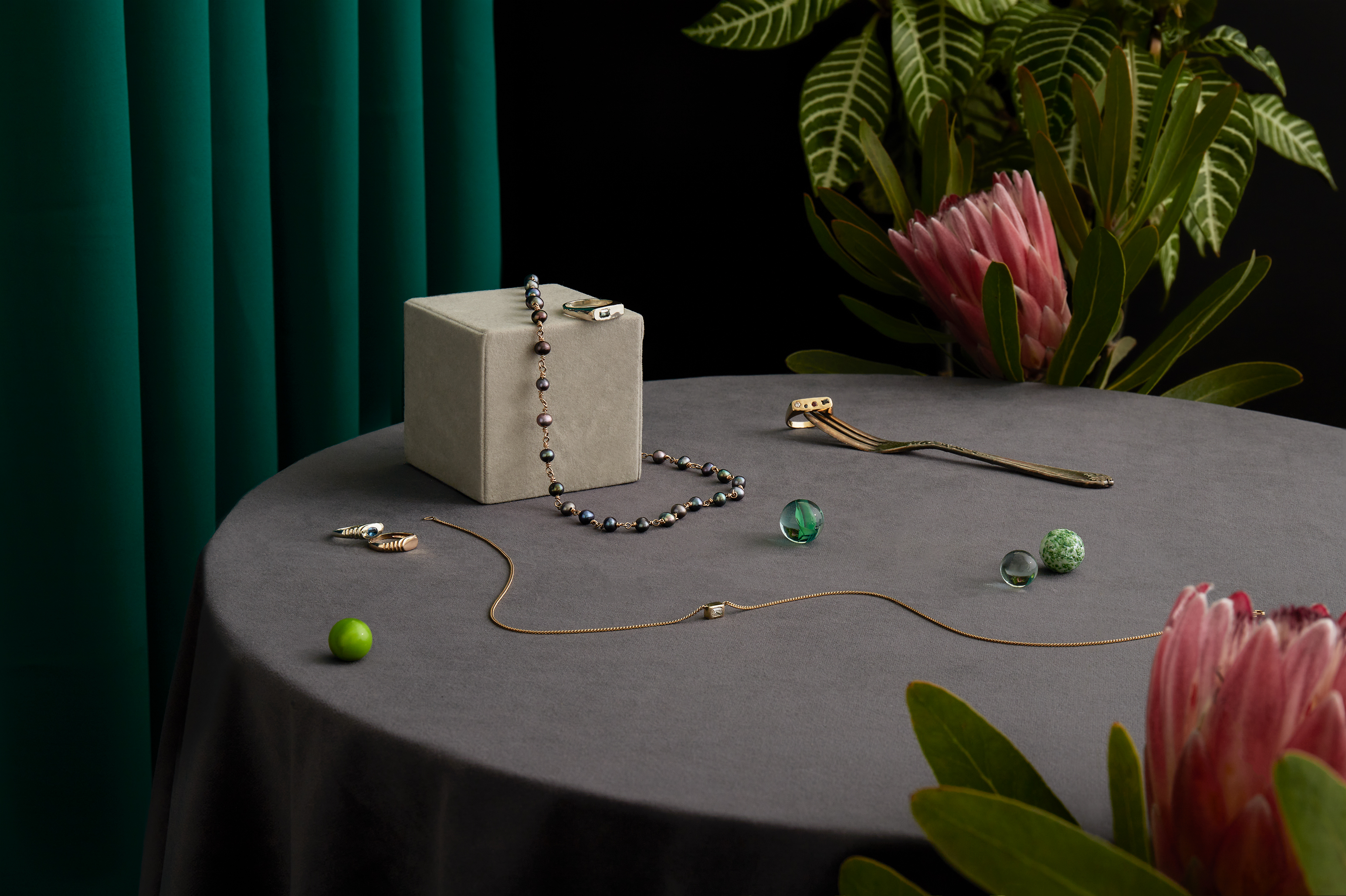 Elegant product shot with jewelry, rings, and gemstones displayed on a grey round table with a gray cube, pink flowers, and green plants.