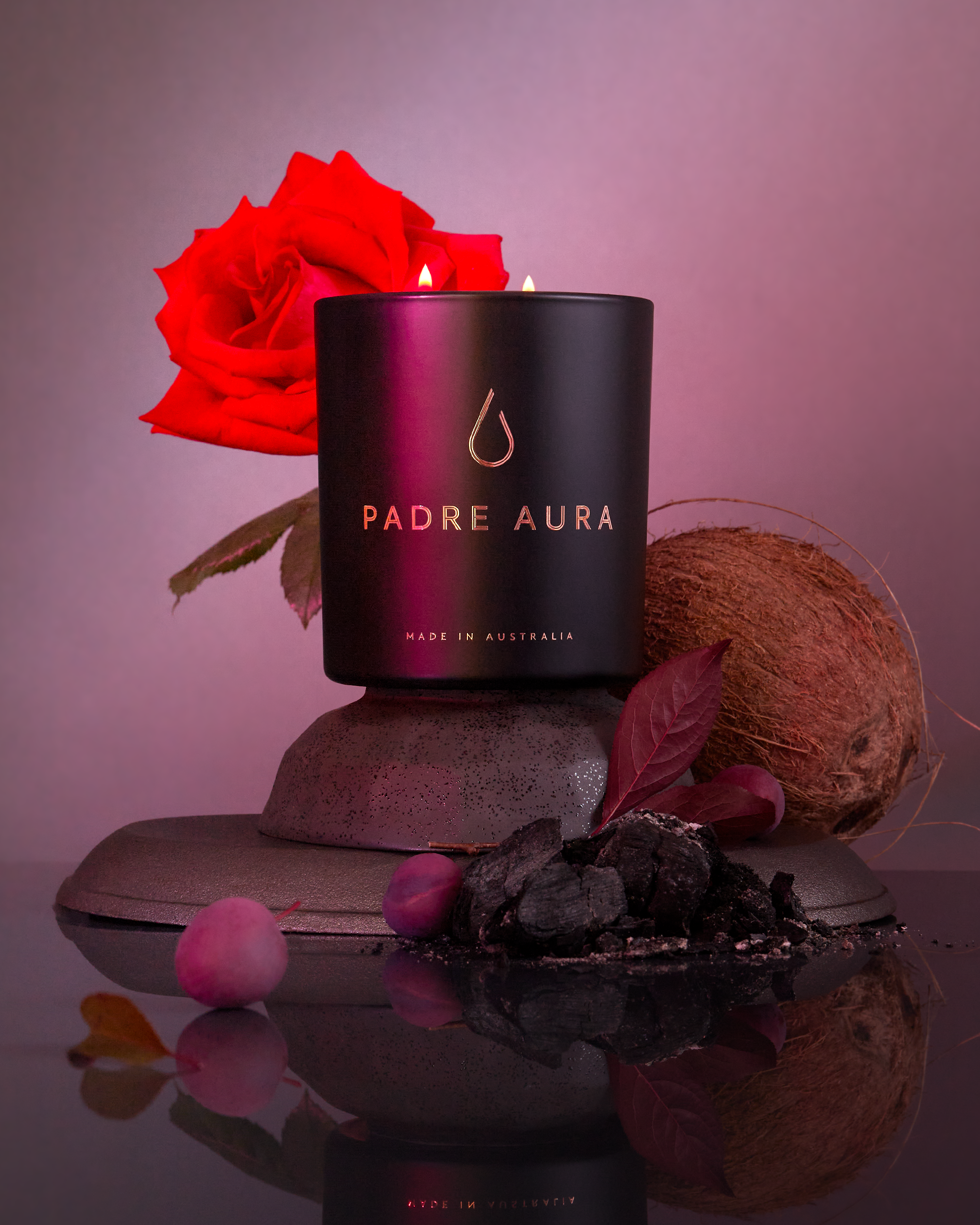Close-up studio shot of a black candle with a red rose, coconut, and charcoal, all reflected on a glossy black surface.
