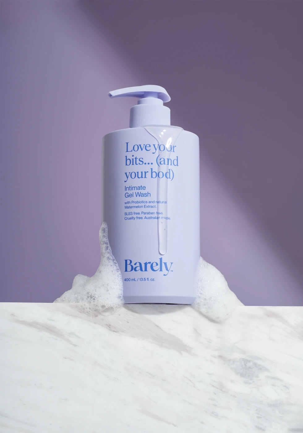 Lavender pump bottle of Barely Intimate Gel Wash resting on a white marble ledge against a purple backdrop with soapy foam and dripping gel.