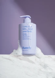 Lavender pump bottle of Barely Intimate Gel Wash resting on a white marble ledge against a purple backdrop with soapy foam and dripping gel.