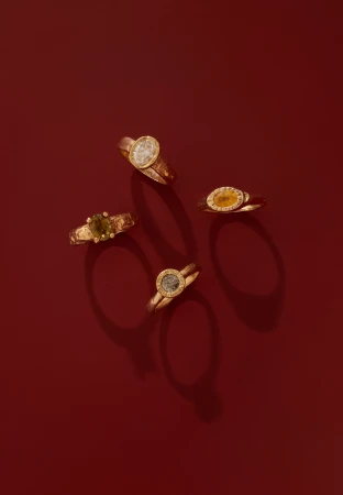 Four gold gemstone rings arranged on a deep red surface with distinct shadows cast beneath each piece.