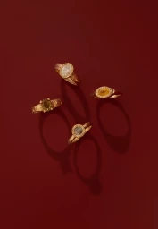 Four gold gemstone rings arranged on a deep red surface with distinct shadows cast beneath each piece.