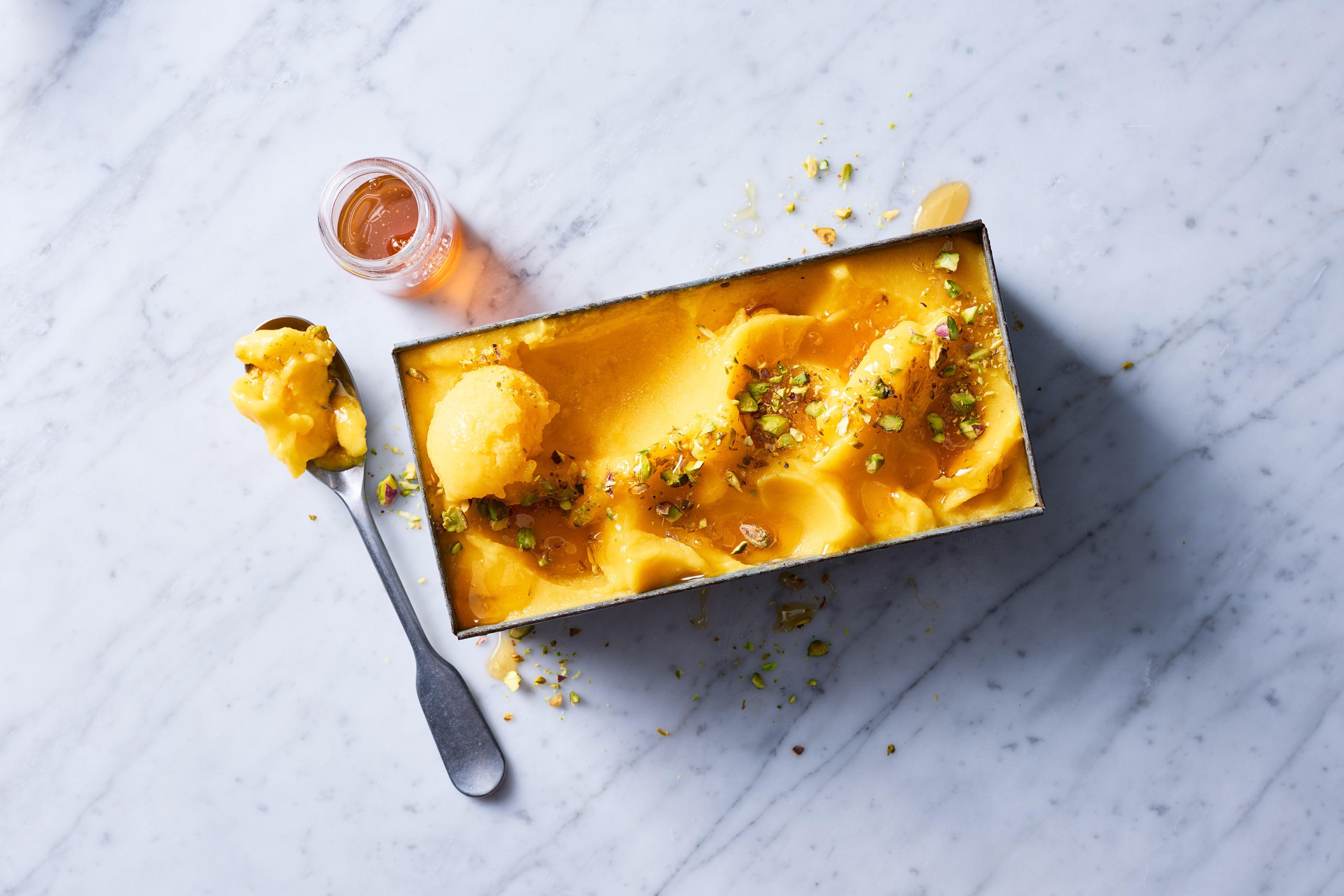 Overhead view of mango sorbet in a metal container with a scoop, garnished with pistachios and honey on a marble surface, studio lighting.