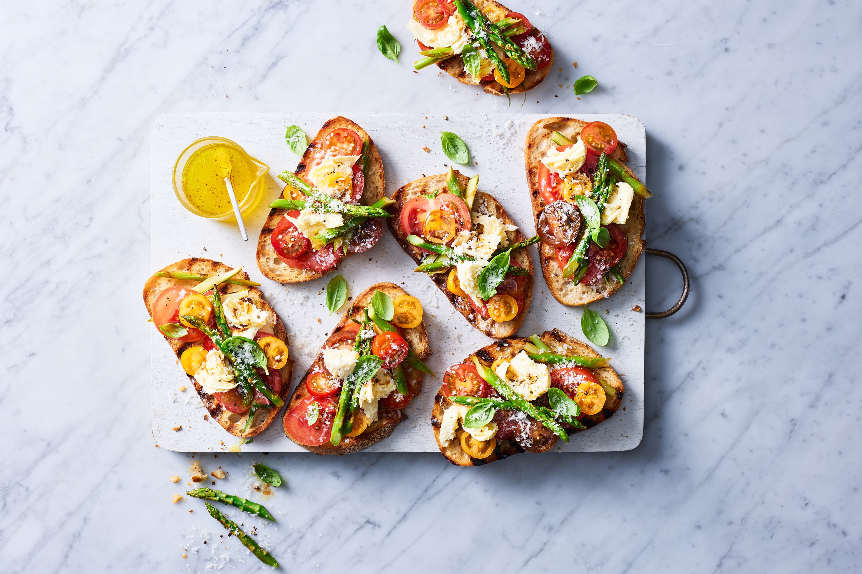 Overhead shot of appetizer toast topped with sliced tomatoes, mozzarella, asparagus, and herbs on a white marble surface.