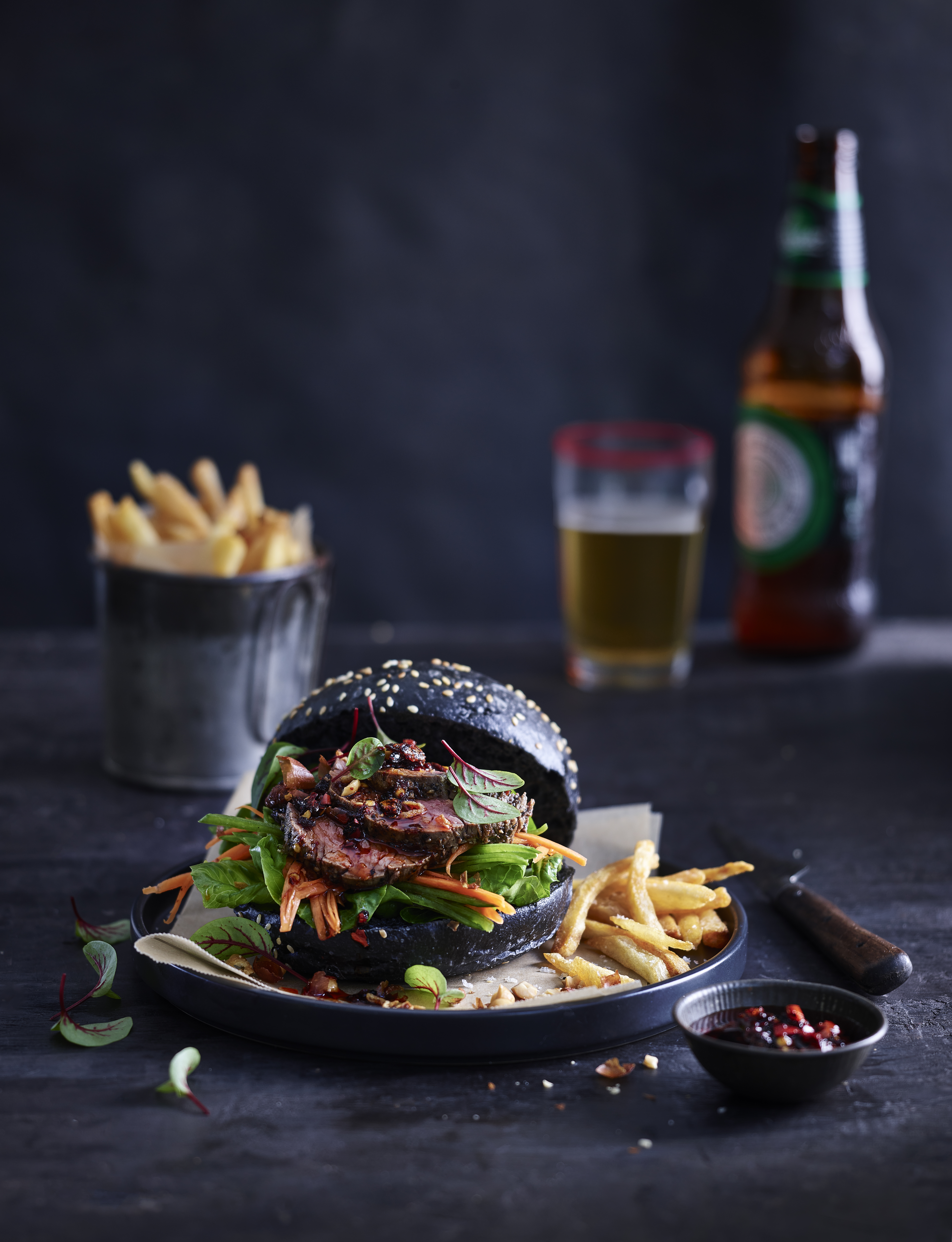 A black bun burger with sliced meat and fresh salad on a plate, accompanied by french fries, beer, and a black surface.