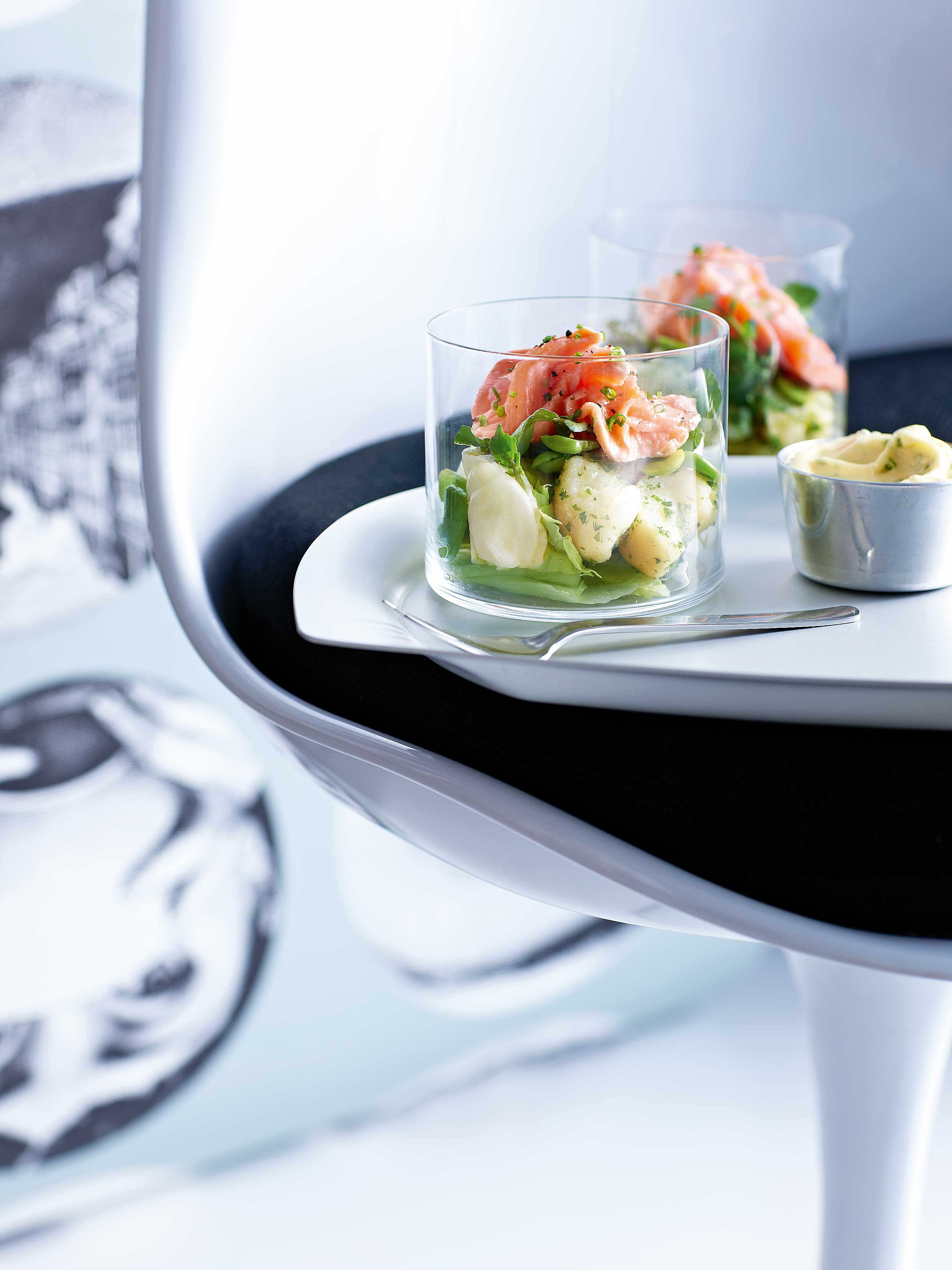 Two glasses of salmon salad with potatoes, lettuce, and greens on a white plate atop a designer serving tray against a light patterned background.