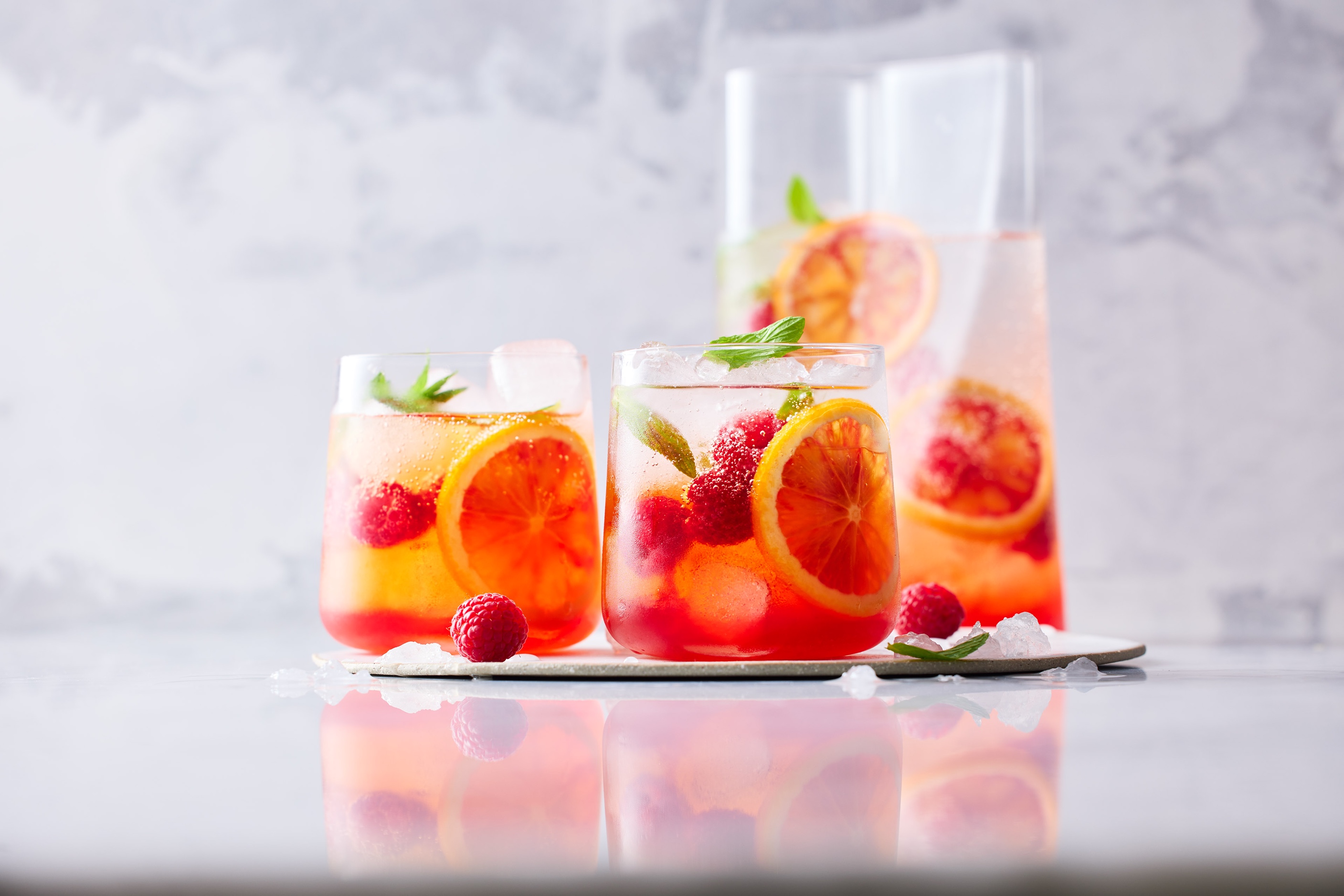 Refreshing fruit-infused water with blood orange slices, raspberries, ice cubes, and mint leaves in clear glasses and a pitcher, indoors.