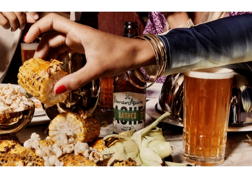 Hand picking up a piece of charred corn from a spread with beer and food styled by Georgina Ropiha