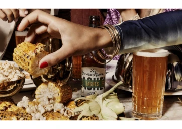 Hand picking up a piece of charred corn from a spread with beer and food styled by Georgina Ropiha