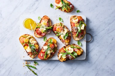 Top-down view of artisan bruschetta with tomatoes, asparagus, and cheese on a white wooden board by Georgina Ropiha