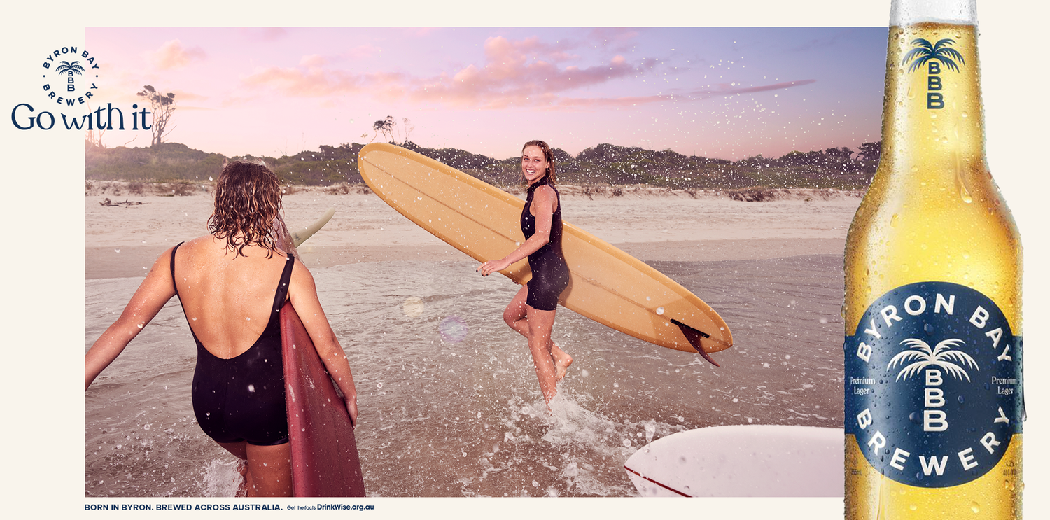 Two women in swimwear walking out of ocean with surfboards; Byron Bay Brewery advertising with bottle visible on right.