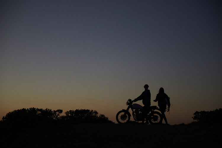 Silhouette of two people and a motorcycle against a dark sunset sky, featuring a rider and a companion on a rural horizon.