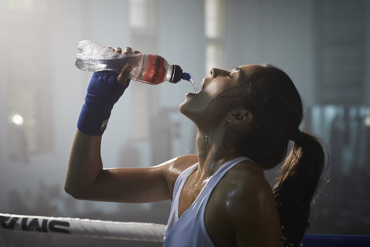 Woman in tank top drinks from water bottle with boxing ring background, wearing blue wrist wraps, perspiring with hair in ponytail.