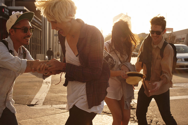 Group of four friends laughing, walking in sunlight in an urban environment. Two men and two women in casual clothes, city background.