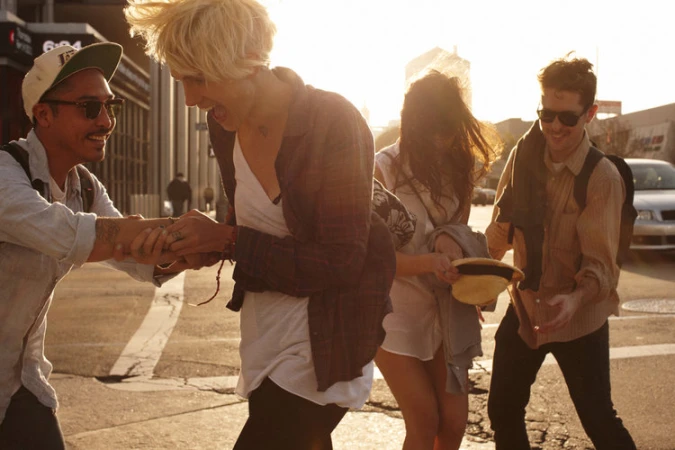 Group of four friends laughing, walking in sunlight in an urban environment. Two men and two women in casual clothes, city background.