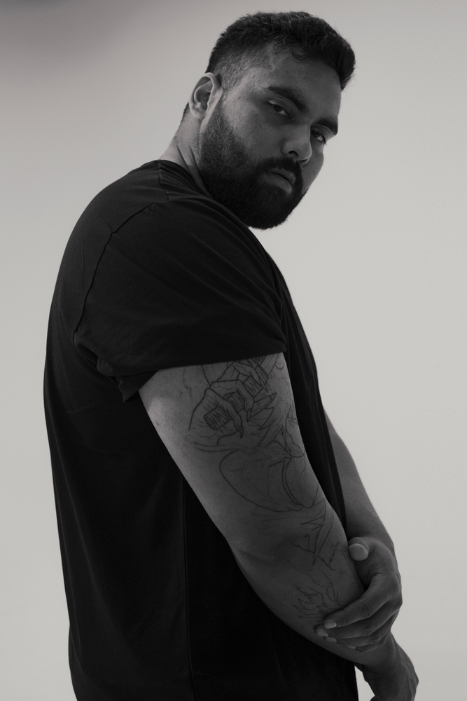 Black and white portrait of a bearded man with tattoos wearing a t-shirt looking at the camera in a photo studio.