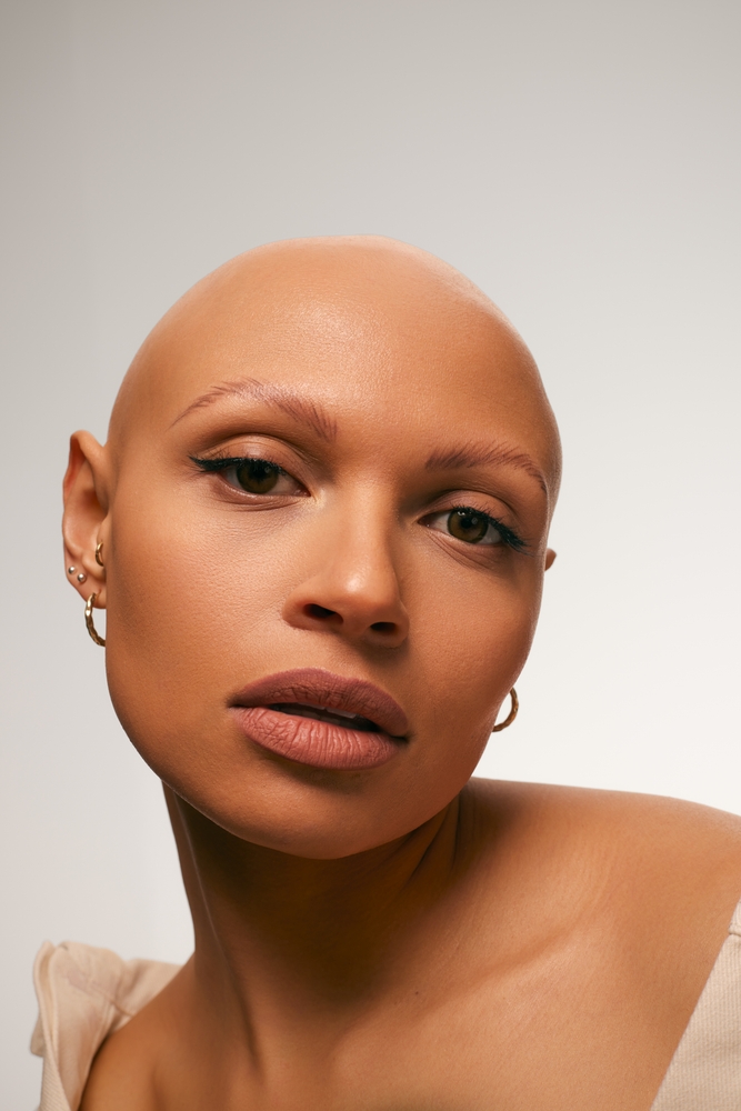 Close-up portrait of a bald woman with light skin, wearing hoop earrings and light-colored clothing, against a plain neutral background.