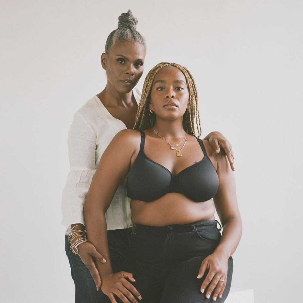 Two women pose in a brightly lit photo studio. The younger woman wears a black bra, while the other wears a white blouse.