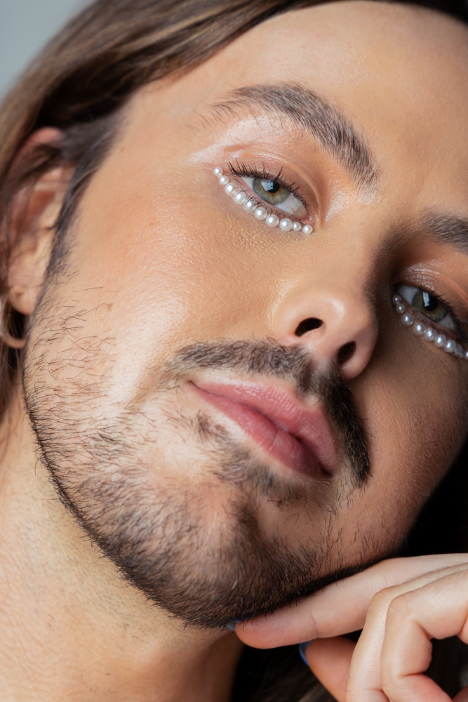 Close-up of a male model with long brown hair and light beard, fair skin, and pearl embellishments around the eyes against a grey background.
