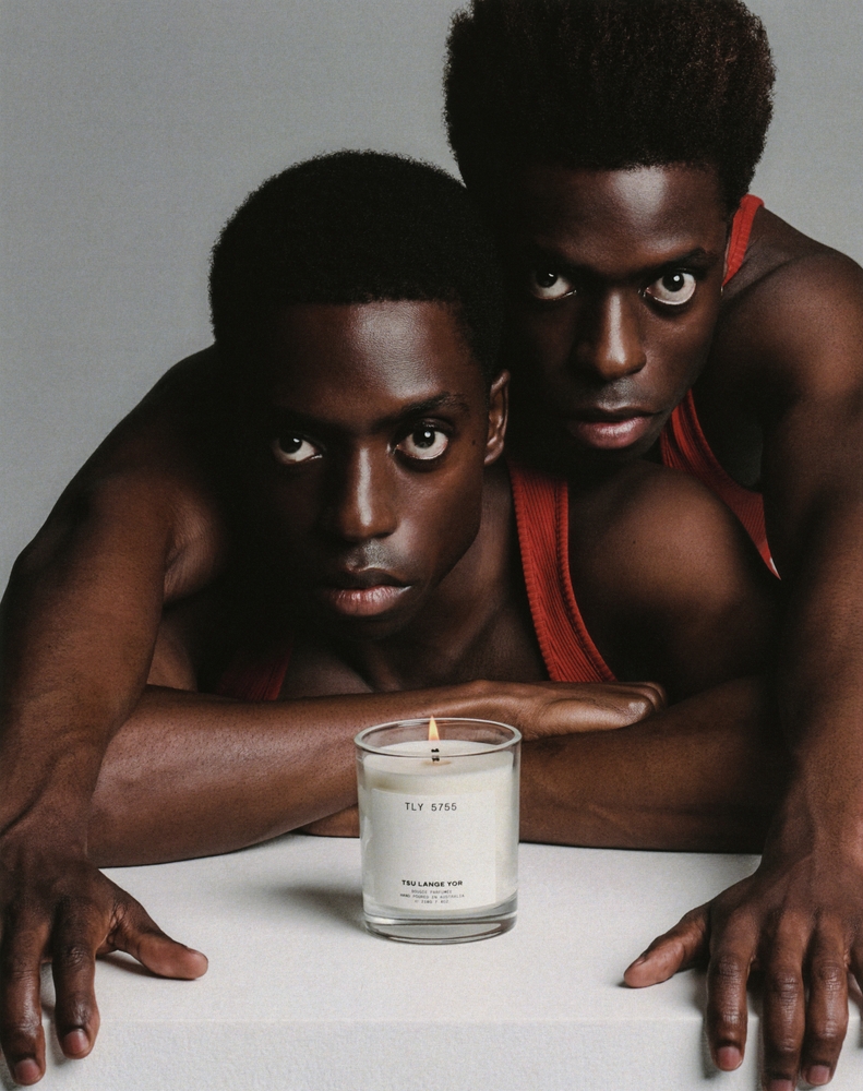 Close-up of two dark-skinned young men in red tank tops behind a lit candle on a white surface against a neutral grey backdrop.