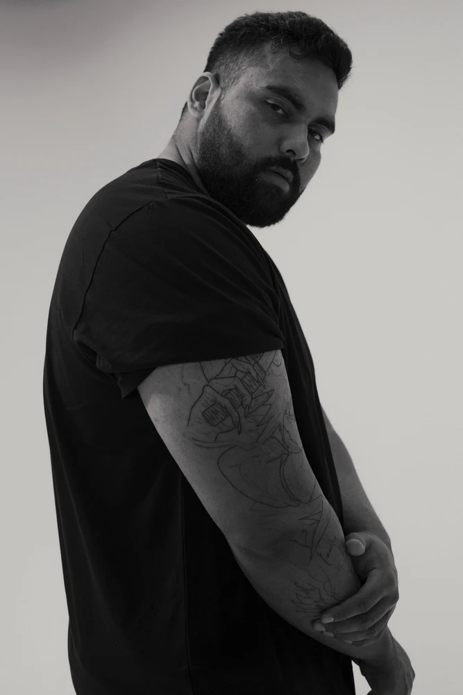 Portrait of a South Asian man wearing a black t-shirt with arm tattoos against a plain white studio background.