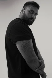 Portrait of a South Asian man wearing a black t-shirt with arm tattoos against a plain white studio background.