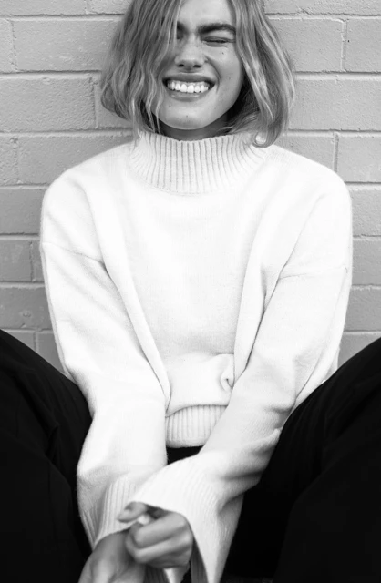 Close-up black and white portrait of a smiling young adult woman with short blond hair wearing a white sweater against a brick wall.