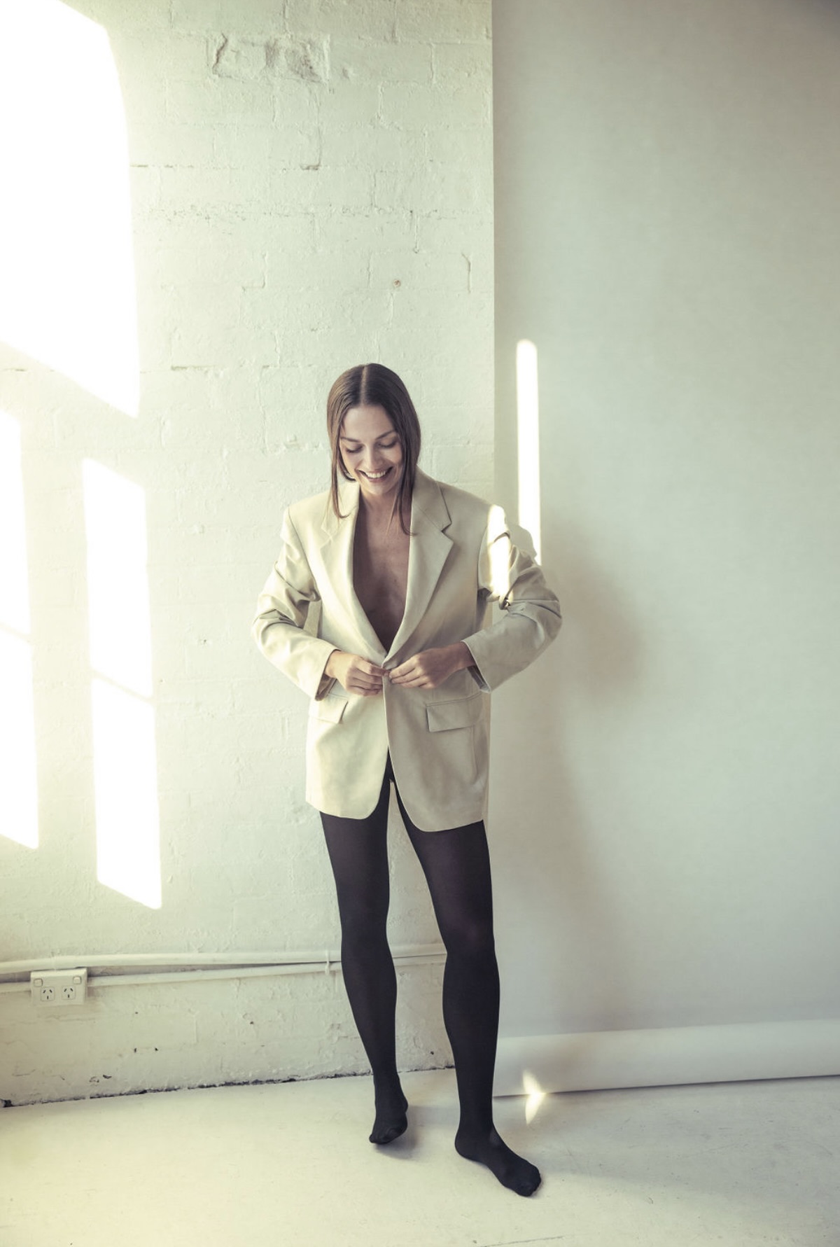 Indoor portrait of an adult woman in a tan blazer and black tights, smiling while standing against a white textured wall.