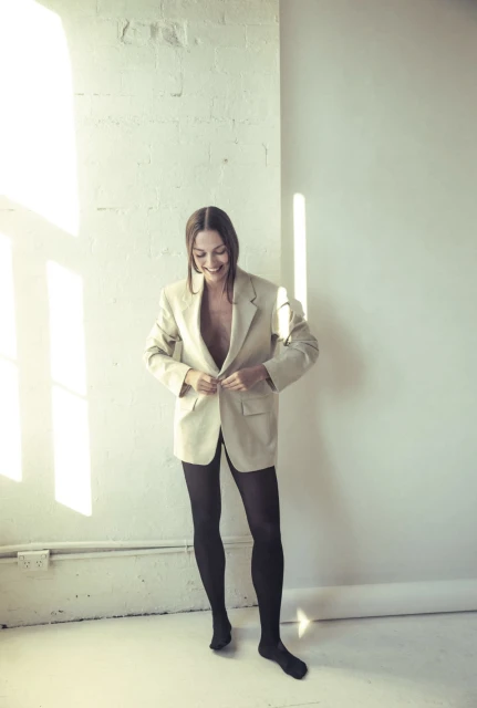 Indoor portrait of an adult woman in a tan blazer and black tights, smiling while standing against a white textured wall.