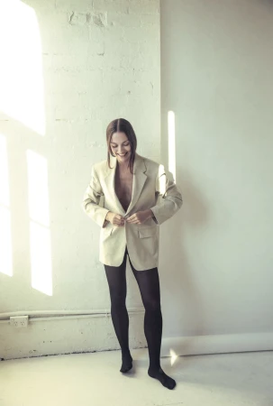 Indoor portrait of an adult woman in a tan blazer and black tights, smiling while standing against a white textured wall.