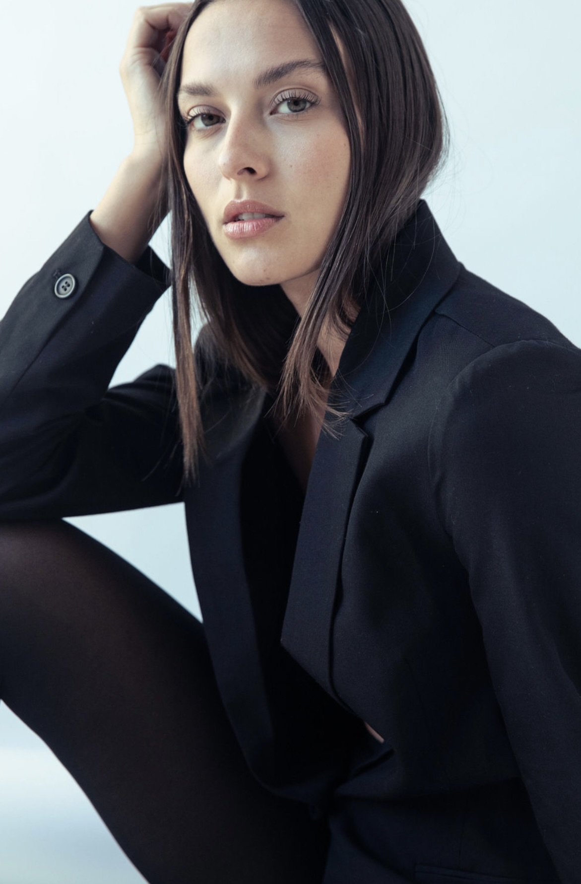 Portrait of a young woman in a black blazer and tights, seated in a photo studio with soft lighting and a neutral background.