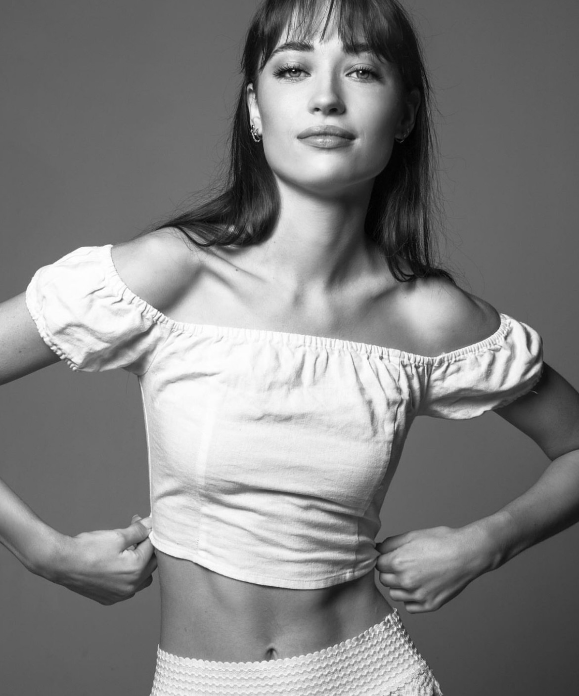 Monochrome portrait of a fair-skinned woman with bangs wearing a white off-shoulder top and shorts against a grey backdrop.