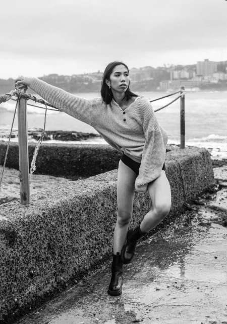 A young woman in a sweater and boots poses on a concrete structure with ocean and urban background in a black and white editorial.