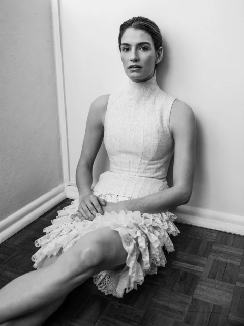 Monochrome portrait of a woman in a textured, high-neck dress with lace details, seated against a white wall on a dark wood floor.