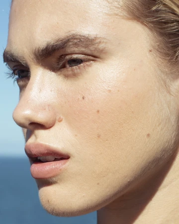 Close-up portrait of a woman's face with natural skin texture, moles, and light makeup under clear sky.