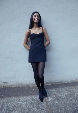 Smiling young woman in a black mini dress and sheer black tights leaning against a light grey wall.