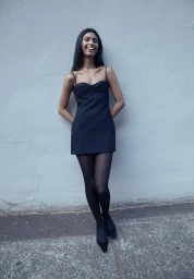 Smiling young woman in a black mini dress and sheer black tights leaning against a light grey wall.