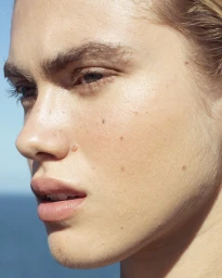 Close-up portrait of a woman's face with natural skin texture, moles, and light makeup under clear sky.